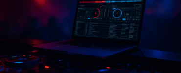 En bærbar computer med Virtual DJ-software åben på skærmen, placeret på et DJ-mixerpult med oplyste knapper og joghjul i rød og blå belysning i et mørkt festlokale.