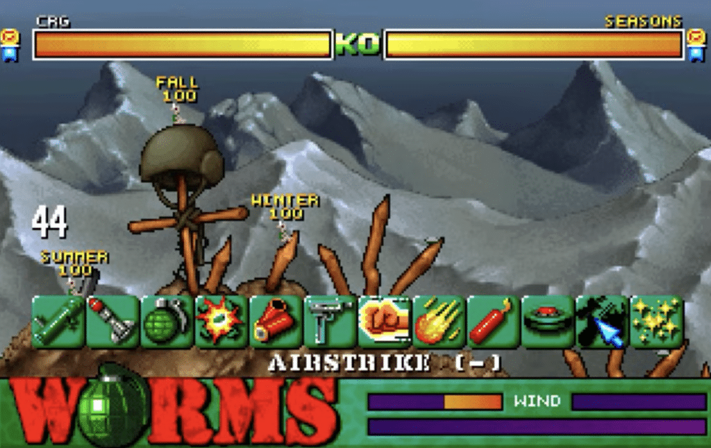 Gameplay fra Worms, hvor flere hold af orme kæmper i et ødelagt bjerglandskab med våbenmenu, vindindikator og sæsonbaseret HUD synlig på skærmen.