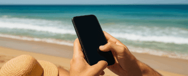 Person spiller offline spil på mobiltelefon på stranden med havet i baggrunden og en solhat ved siden af.