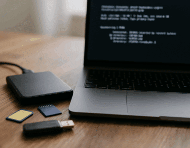 Ekstern harddisk, SD-kort og USB-nøgle på træbord ved siden af bærbar computer med terminalvindue åbent – datagendannelse i gang.