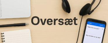 Et skrivebord med oversættelsesværktøjer – en bærbar computer, notesbog, kuglepen, headset og en smartphone med oversættelsesapp – med ordet "Oversæt" skrevet i midten på træoverfladen.