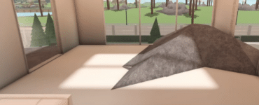 Et screenshot fra Roblox-spillet Welcome to Bloxburg, der viser byggemenuen nederst med kategorier som Cabinets, Comfort, Decorations og Lighting. I baggrunden ses et moderne lyst rum med store glaspartier, en minimalistisk lysekrone og udsigt til palmer og et grønt landskab.